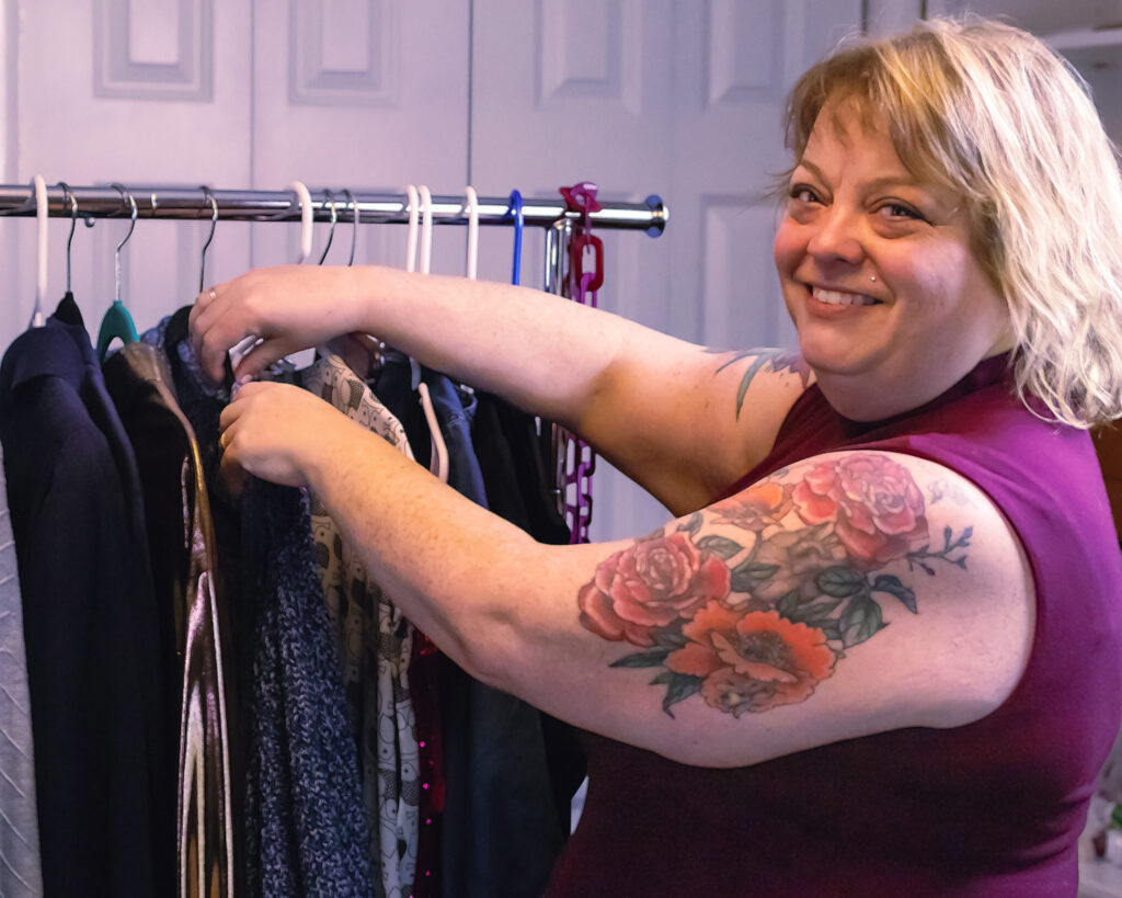 A plus size woman with a large tattoo on her arm stands at a rack of clothing, smiling toward the camera.